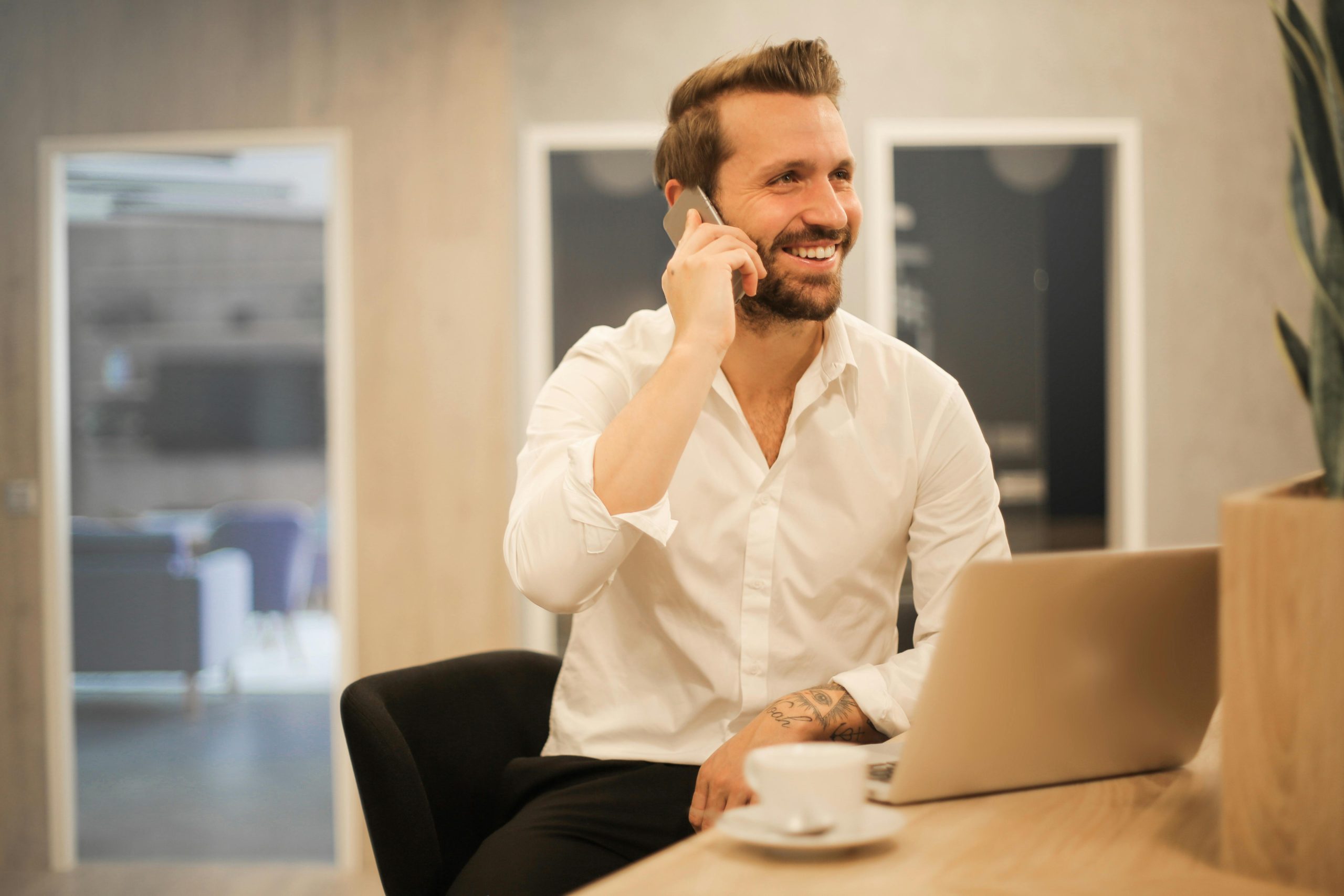 Homme souriant en chemise blanche parlant au téléphone, assis devant un ordinateur portable dans un bureau moderne.
